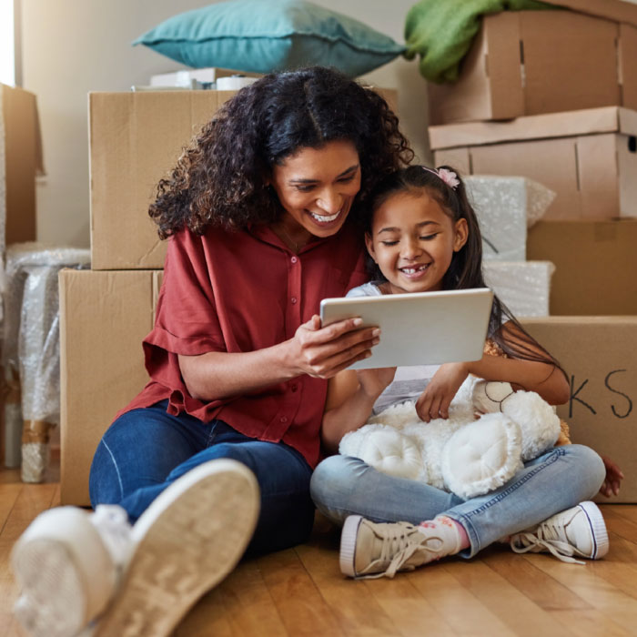 Mom sitting on the floor in front of moving boxes with daughter laughing at something on a tablet